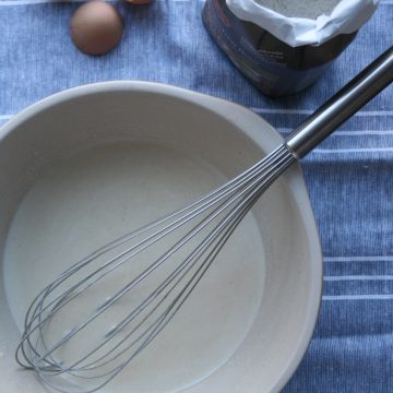 pancake batter in a bowl with a whisk and flour bag above.