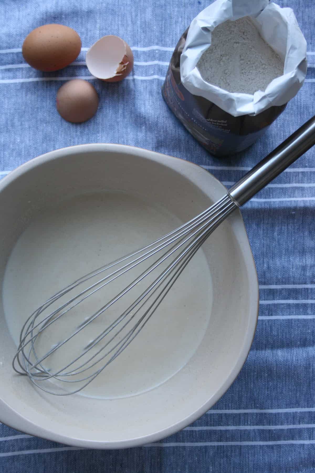 pancake batter in a bowl with a whisk and flour bag above.