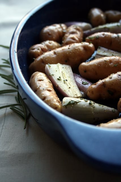 Potatoes and shallots dressed in a blue baking dish with rosemary to the side.