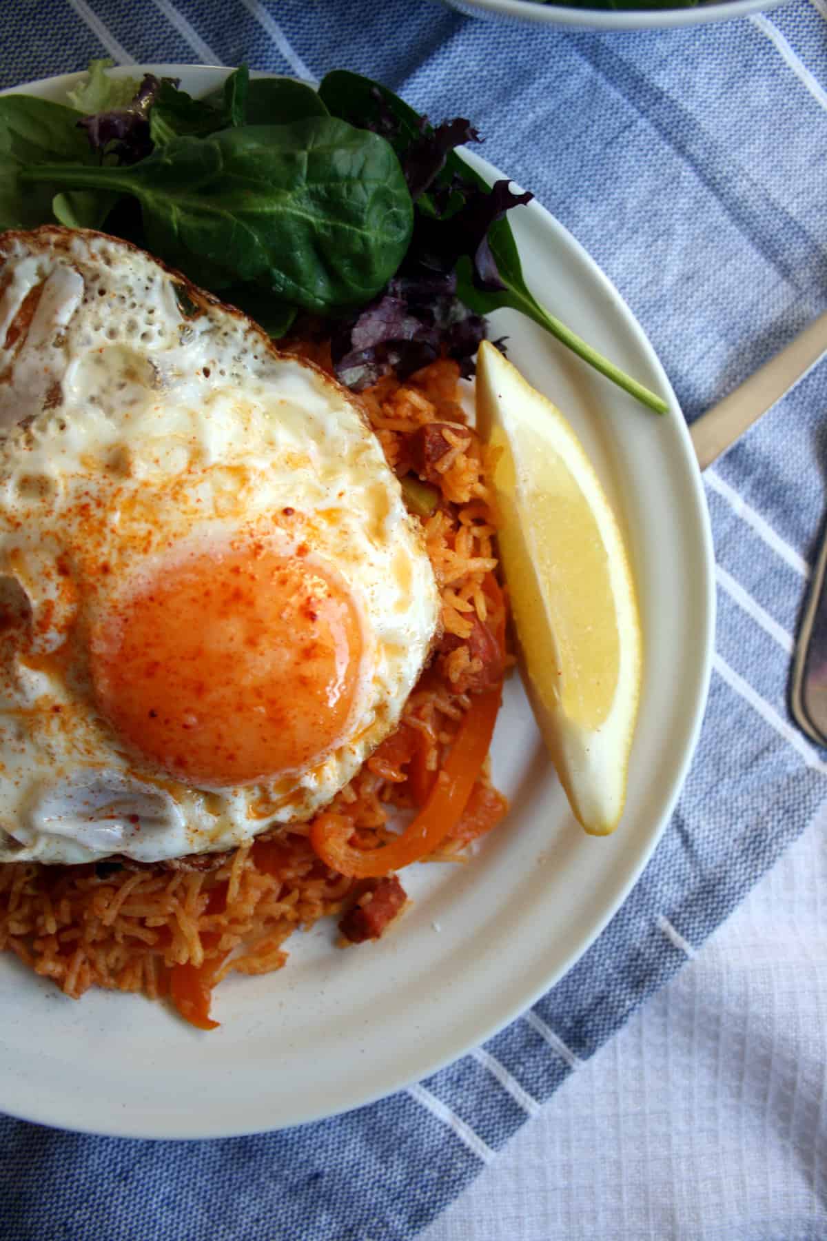 A plate of paella rice and egg on top with salad and a lemon wedge. 