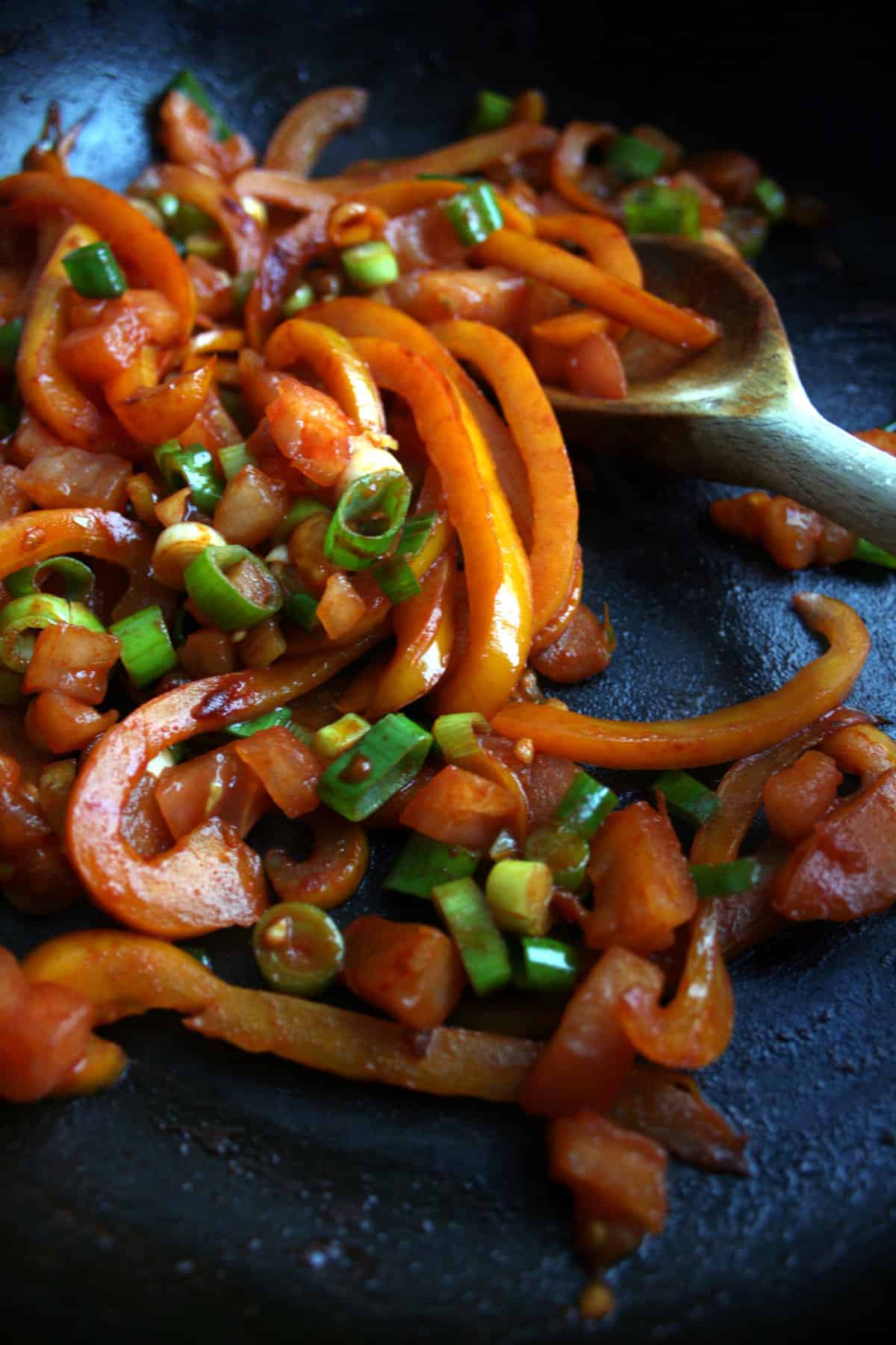 A skillet with cooked peppers, green onions, tomatoes and a wooden spoon.