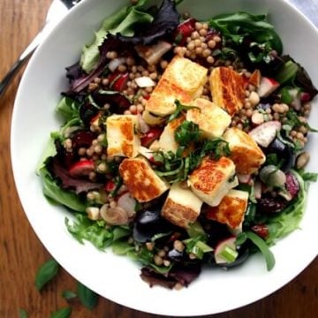 A bowl of crispy halloumi on top of cherries with maftoul and leaves.