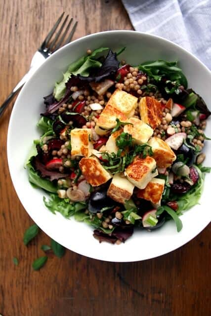 A bowl of crispy halloumi on top of cherries with maftoul and leaves.