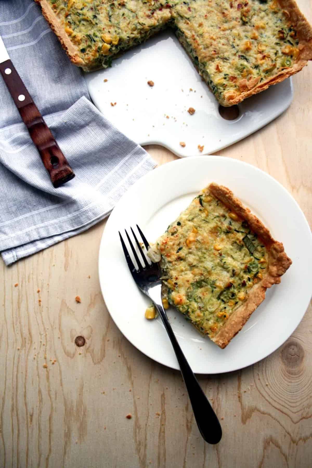 A plate with a slice of tart and a fork with napkin and cutting board above.