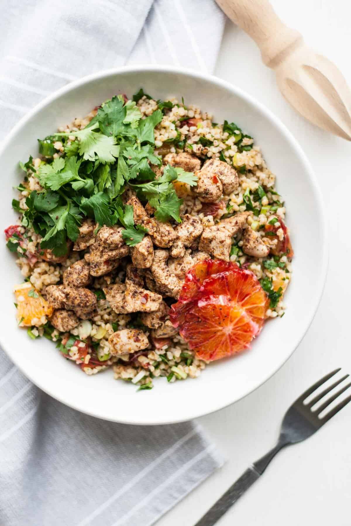 A bowl of chicken tabbouleh with slices of orange and herbs with a fork to the side.