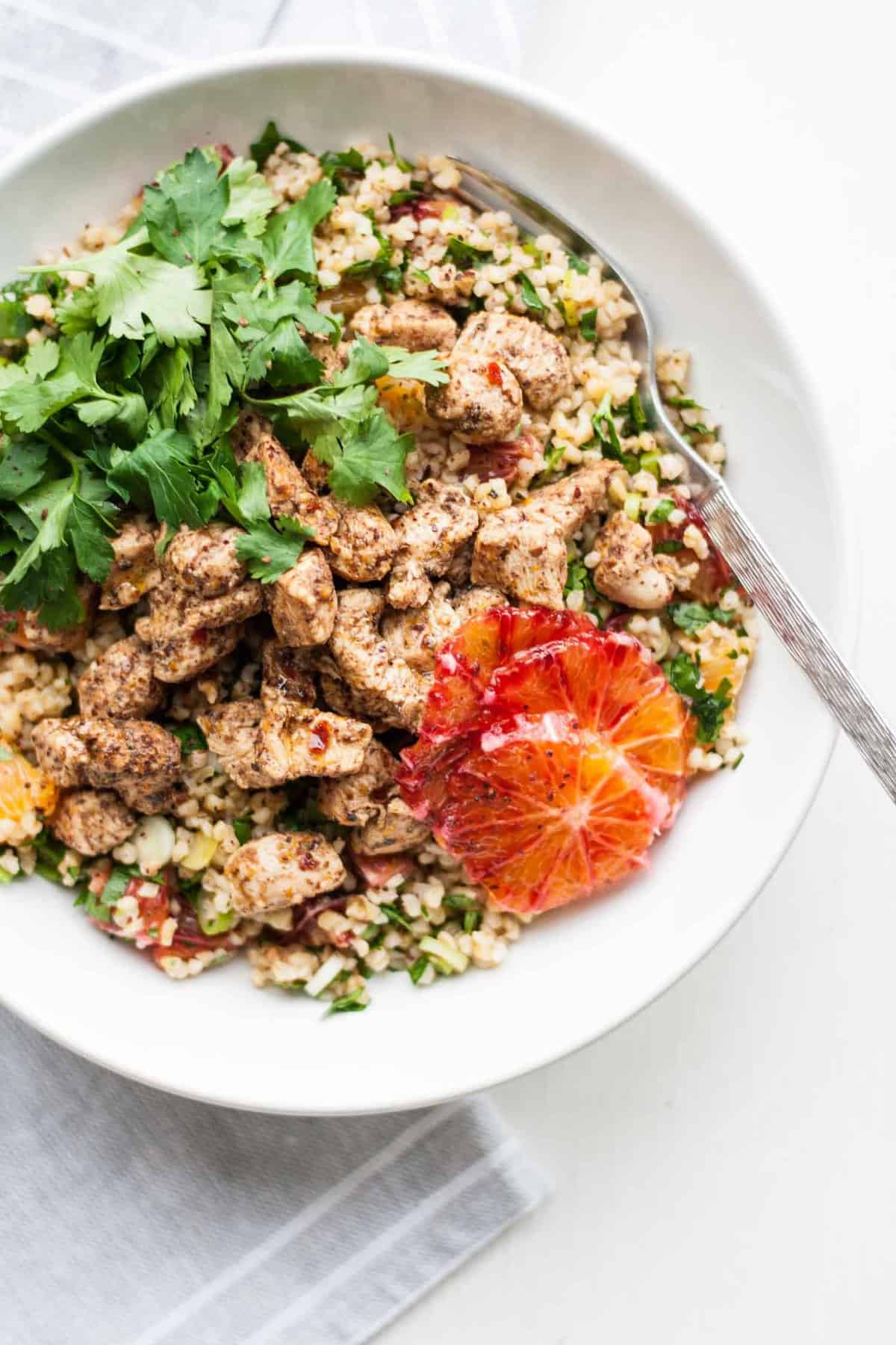 A bowl with chicken, tabbouleh, herbs and slices of orange.