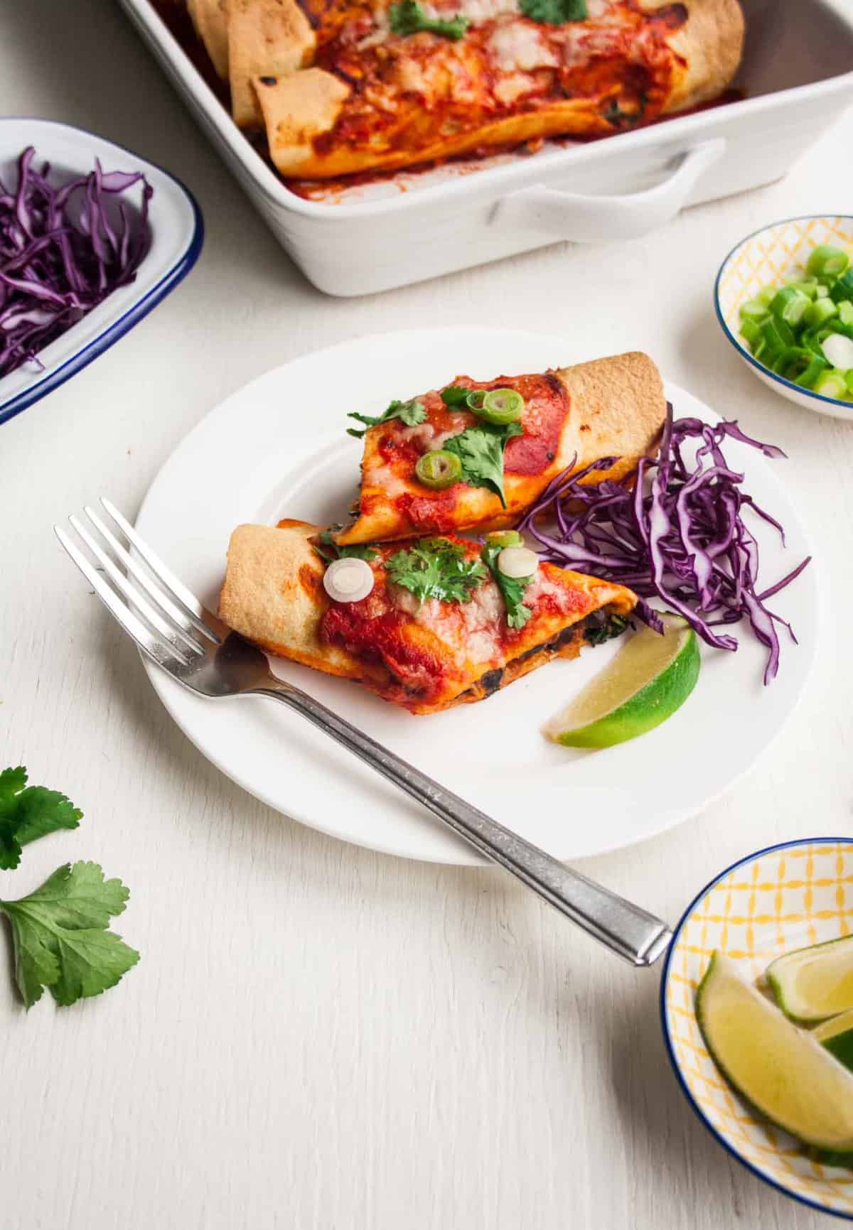 A plate with and cut in half enchilada with a fork, pickled cabbage and lime.