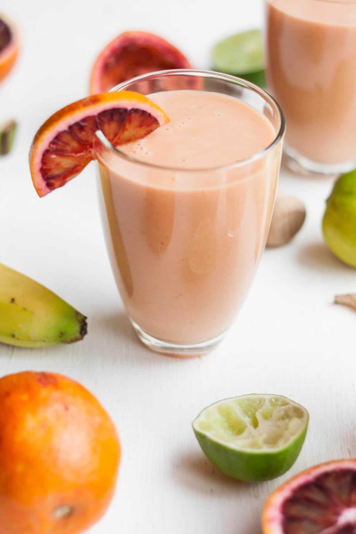 A glass of orange smoothie with lime, orange and banana around.