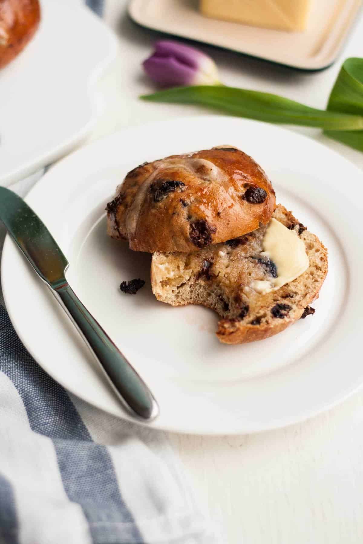 A plate with a cut open hot cross bun with butter on it and a bite taken.