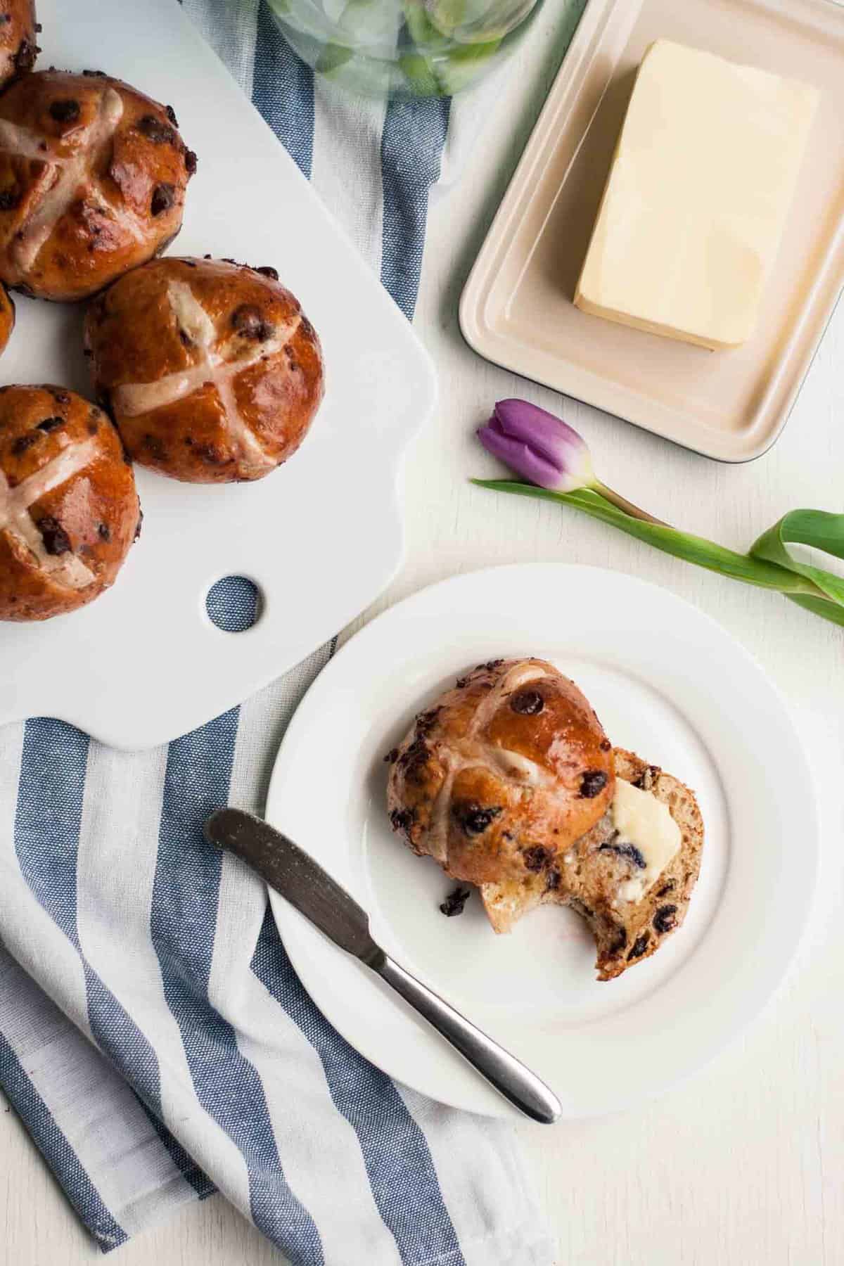 A plate and board with hot cross buns on with one spread with butter and a bite taken.