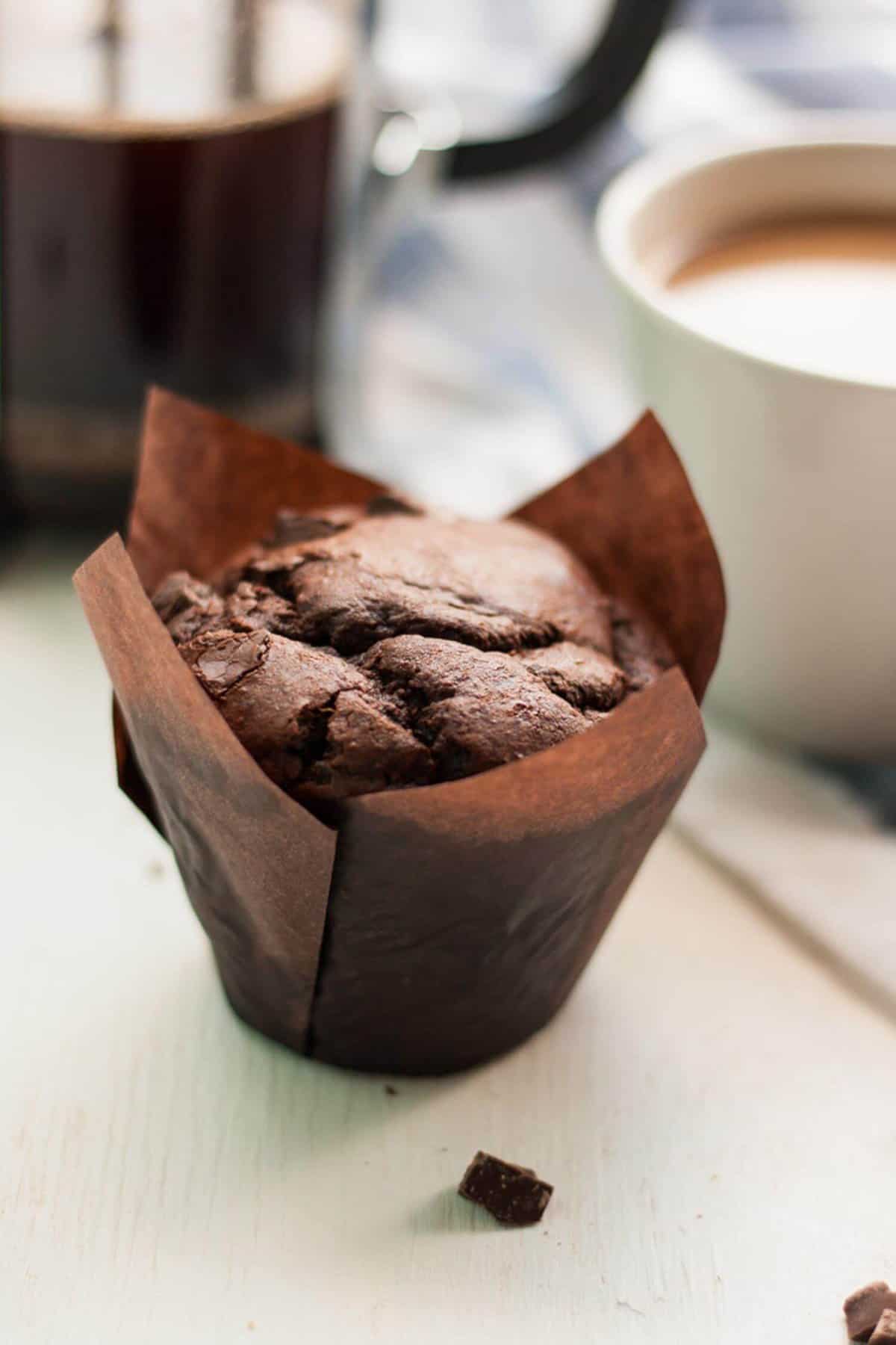 A cup of coffee behind a single chocolate chip muffin in a tulip case.