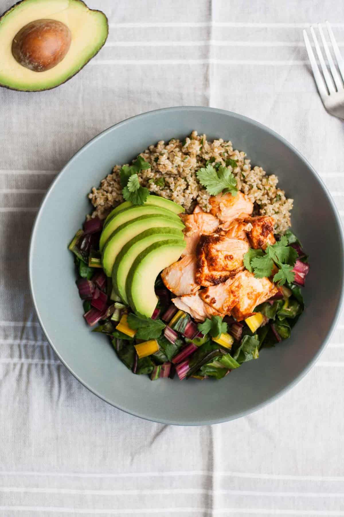 A bowl of grains, salmon and chard with avocado on top.