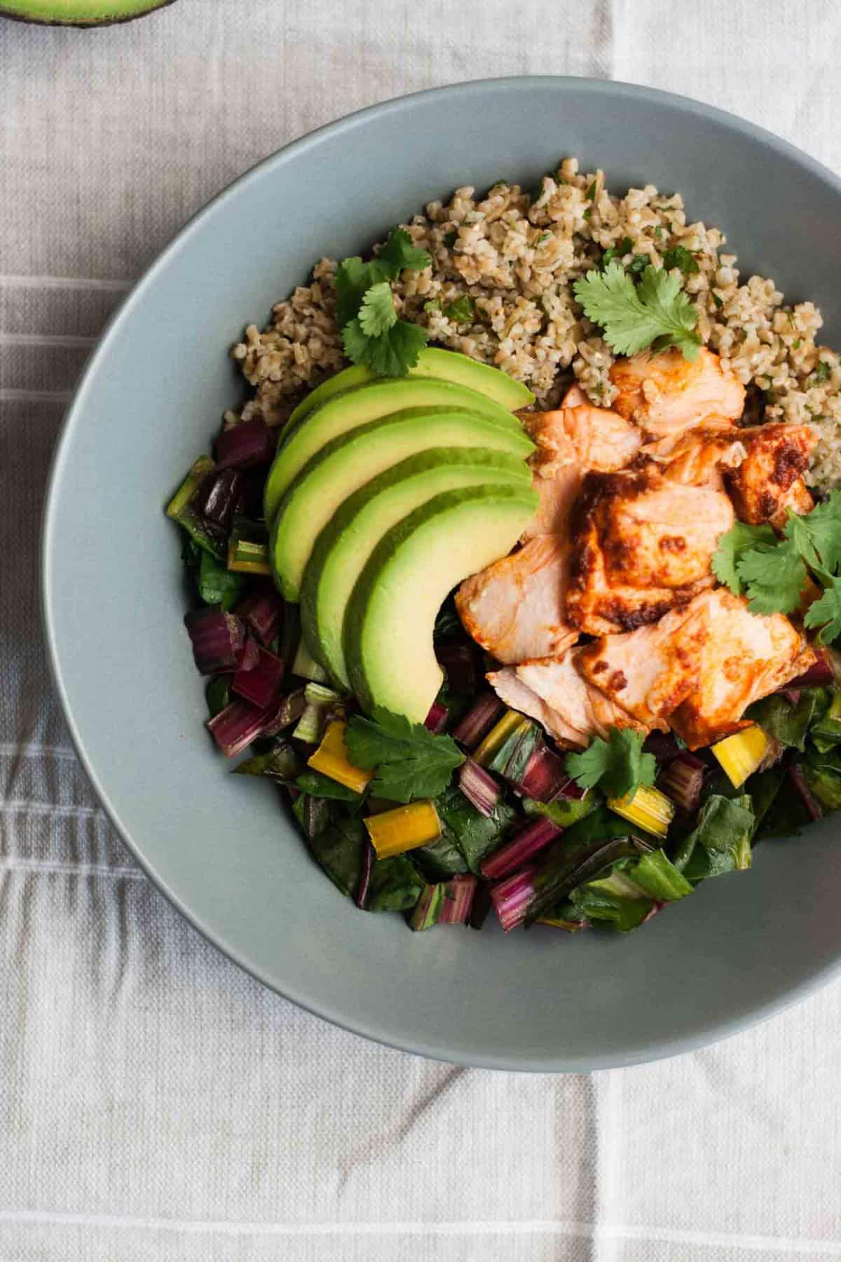 A bowl of flaked salmon on top of chard, grains and with avocado.