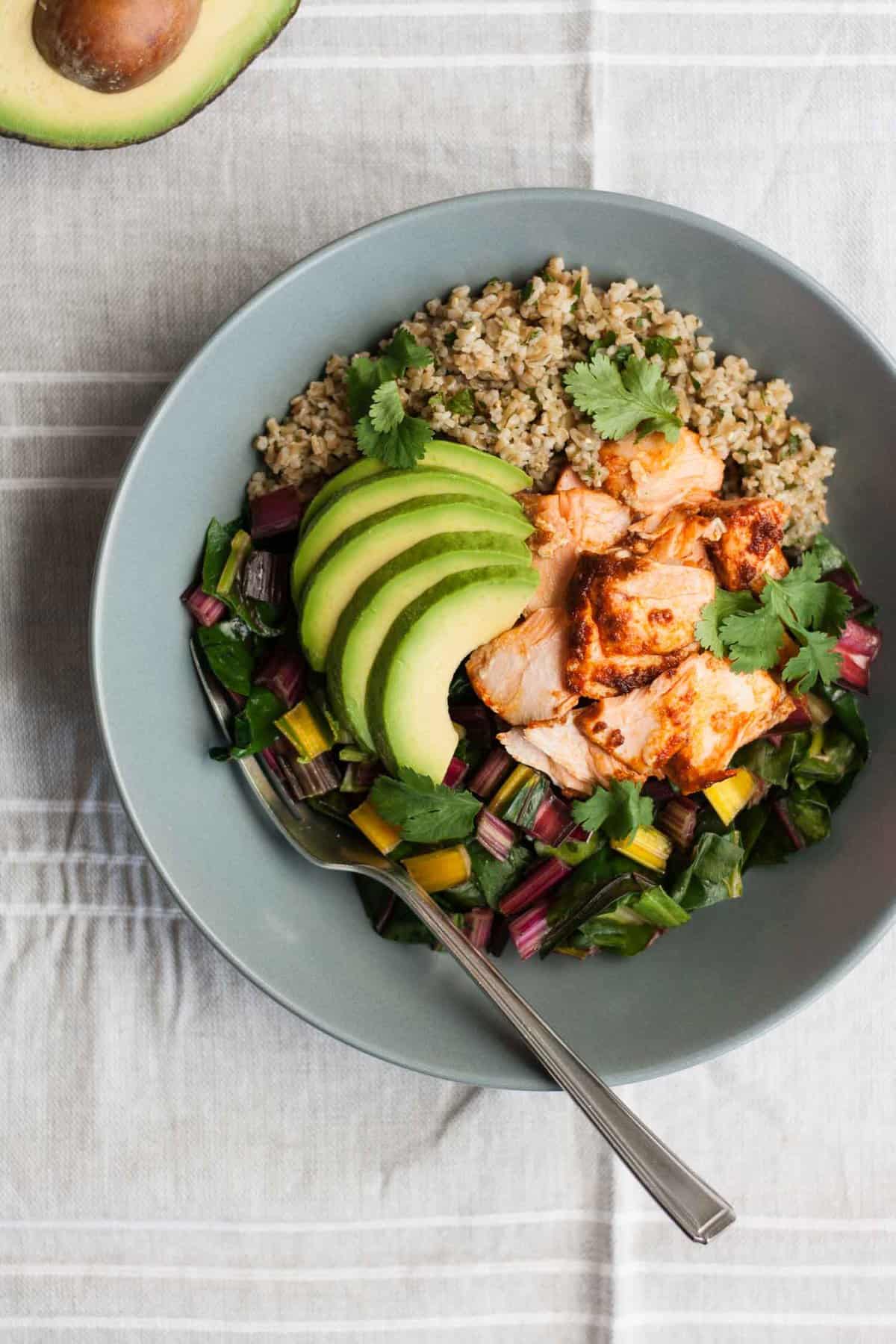 A bowl of grain with chard, flaked salmon avocado and a fork.