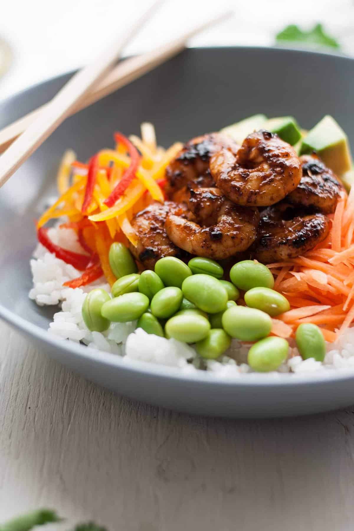 Spicy prawns on top of veggies and rice in a bowl with chopsticks.