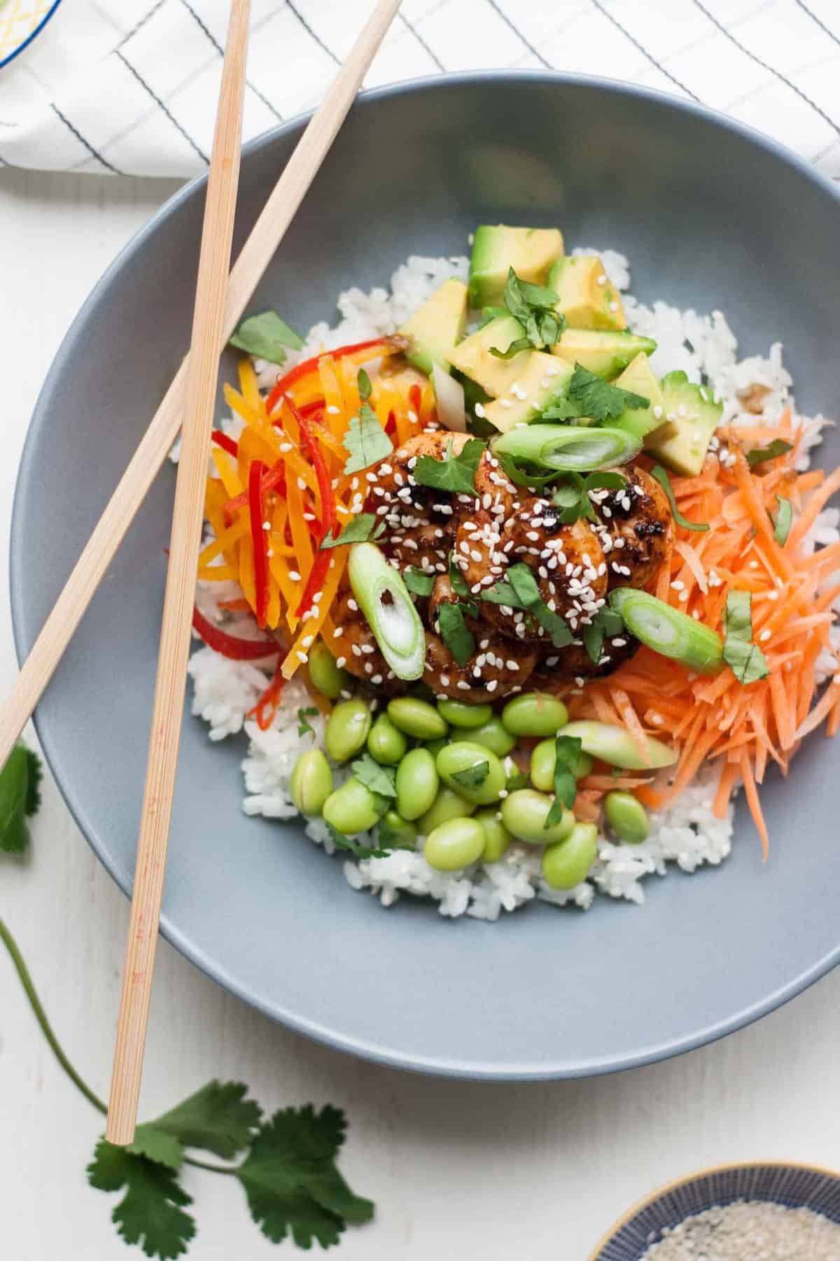 Spicy sticky prawns served with salad items on top of rice in a bowl with chopsticks.