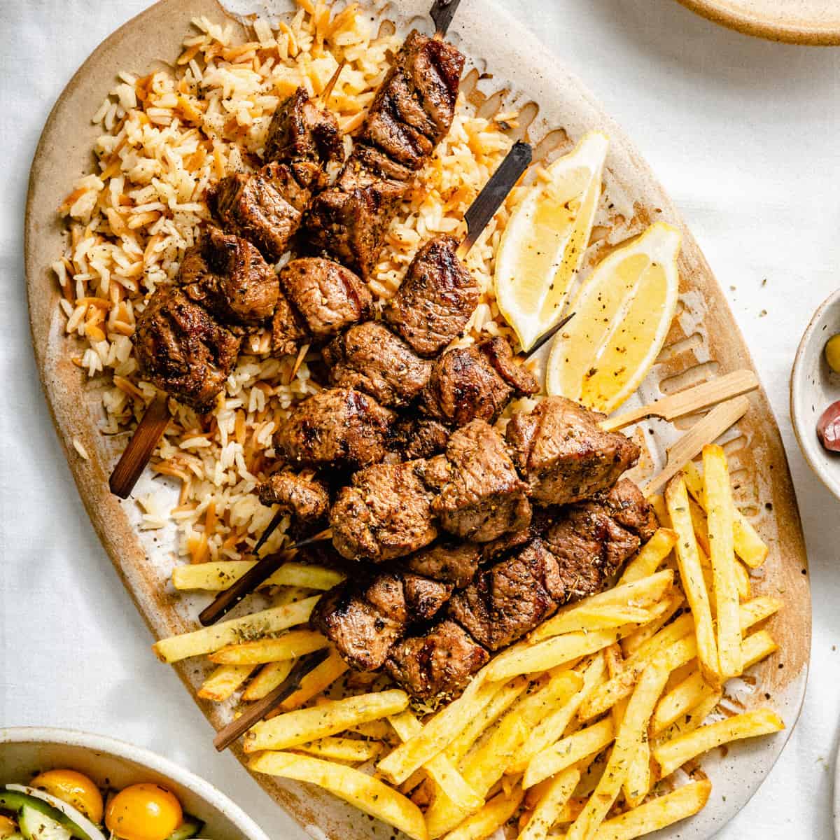 A platter with chargrilled Greek lamb souvlaki, orzo rice, fries and lemon wedges.