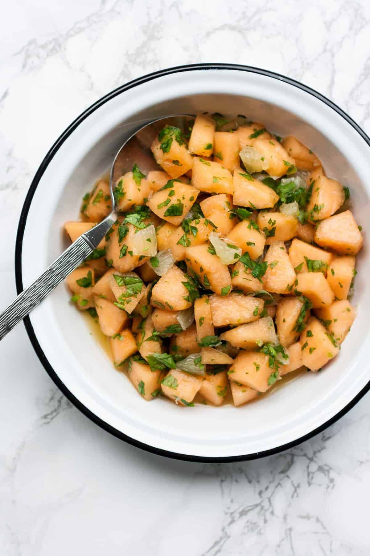 A bowl of melon cubes dressed with lime and herbs with a spoon.