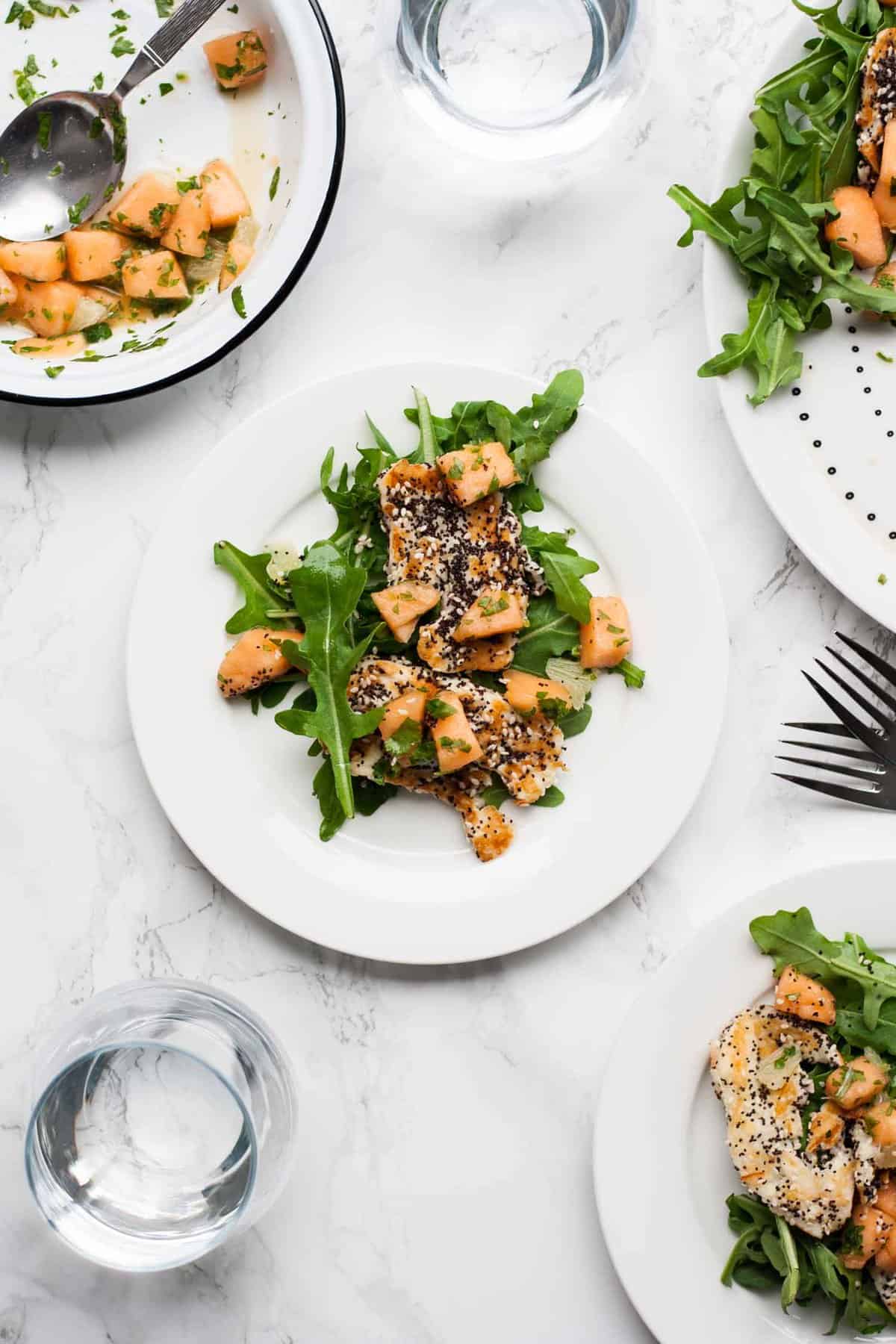 A plate of melon, halloumi salad on a marble work surface with forks and water.