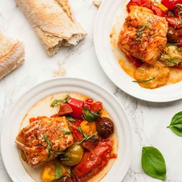 Two plates of baked cod with tomatoes and a broken open baguette to the side.