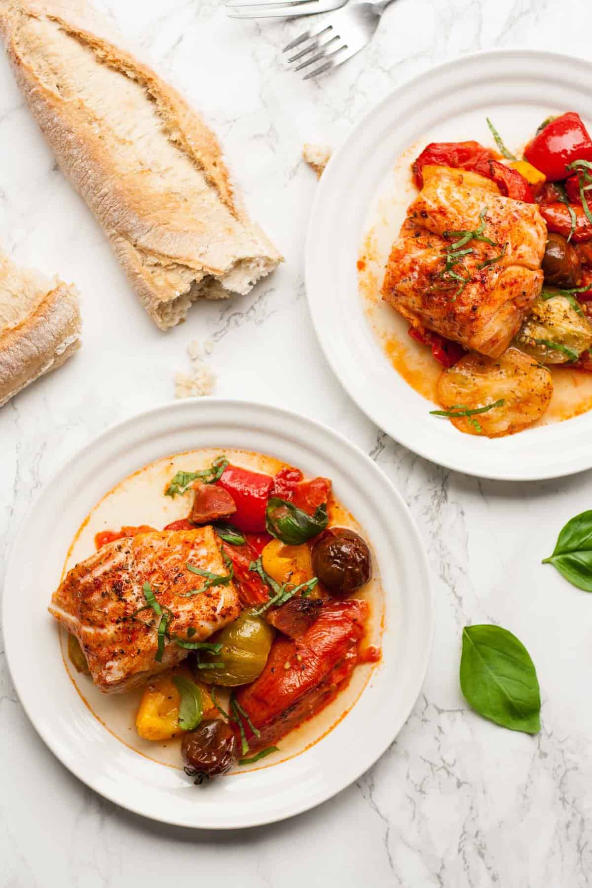 Two plates of baked cod with tomatoes and a broken open baguette to the side.