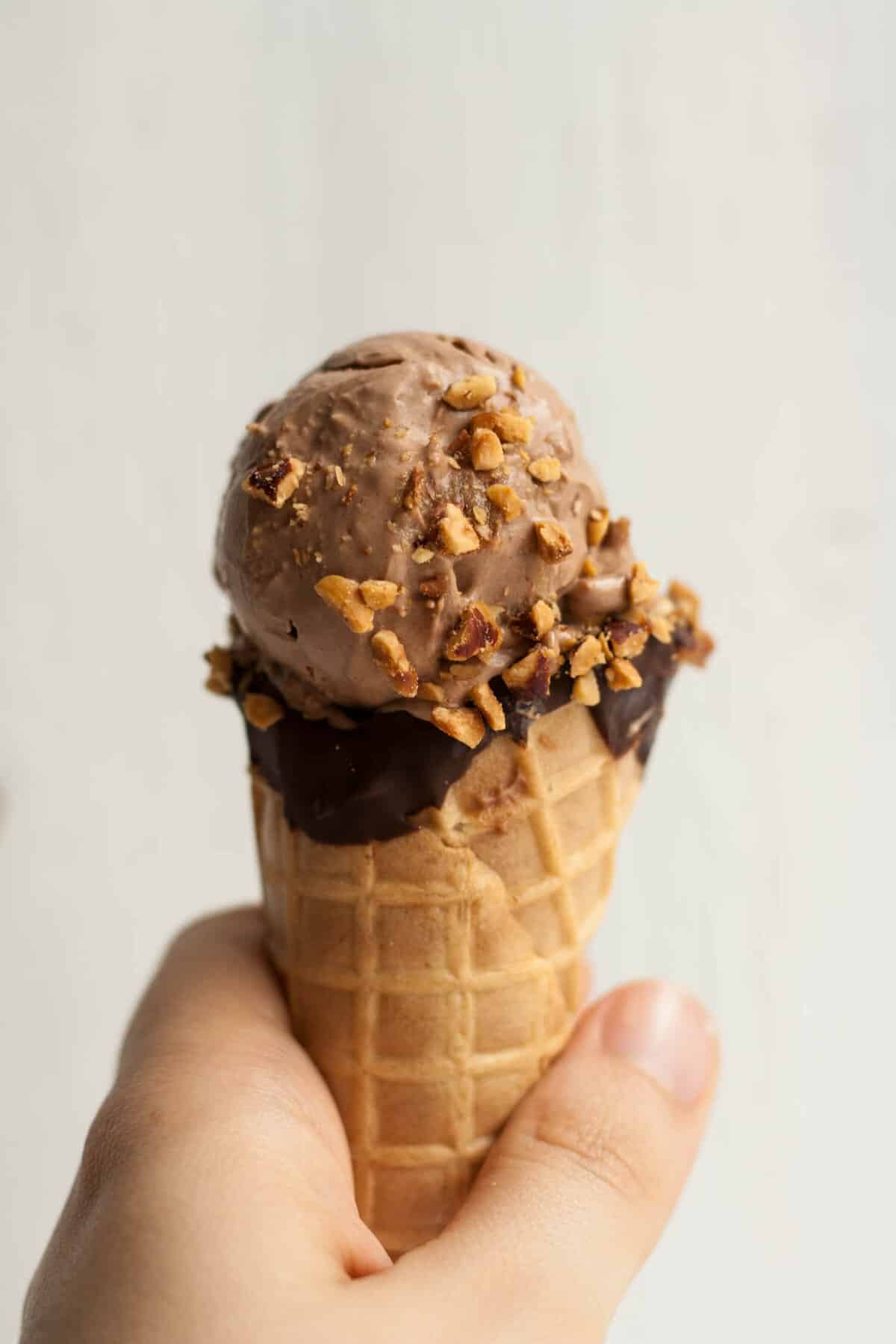 A person holding a mocha ice cream waffle cone with nuts sprinkled on top.