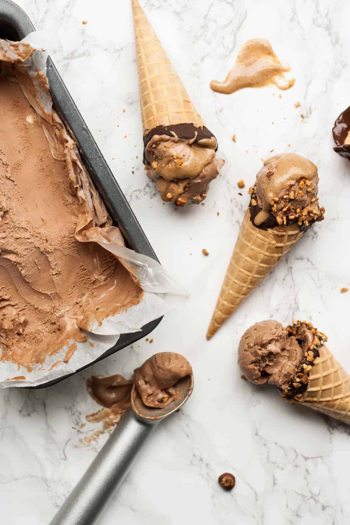 Multiple cones of ice cream on a marble surface with the tin of mocha ice cream and scoop to the side. 