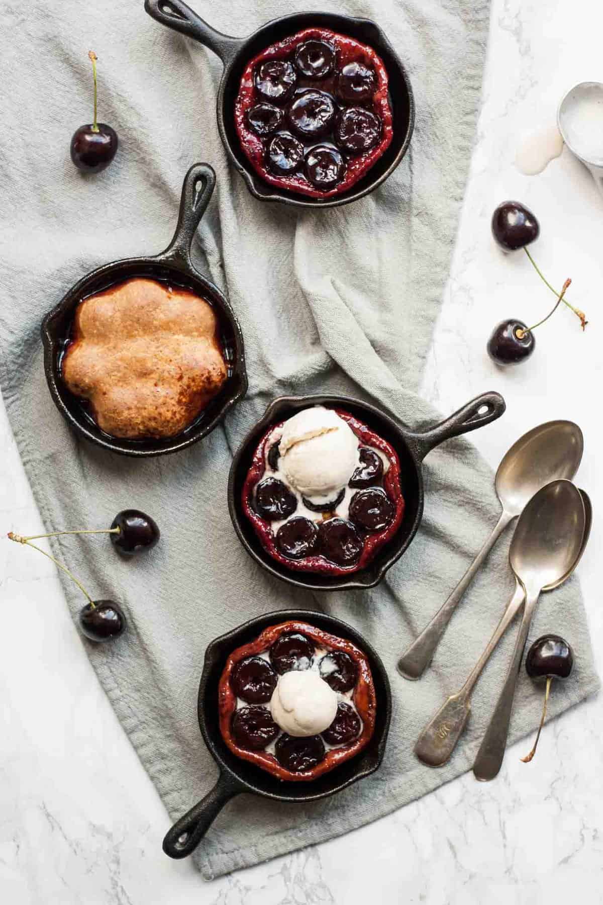 A napkin on a table with mini skillets containing cherry tarte tatin with spoons and cherries around.