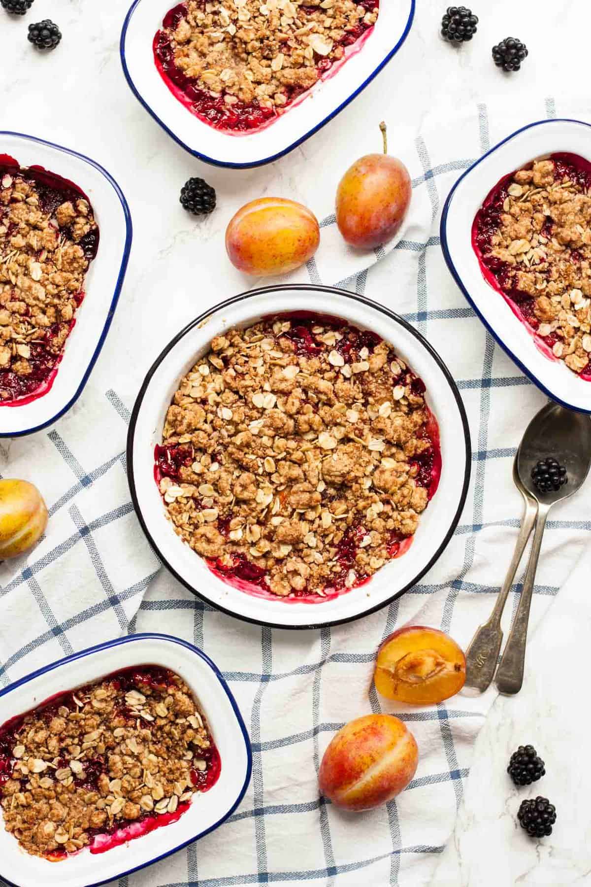 An array of plum and blackberry crisp dishes on a napkin with fruit around.