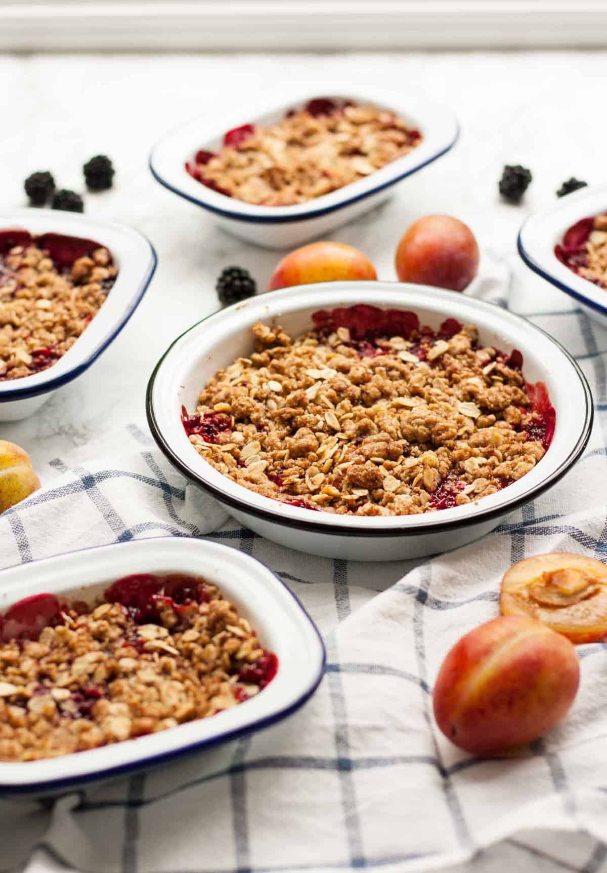 Multiple baking dishes with blackberry and plum crisp inside and plums and blackberries around.