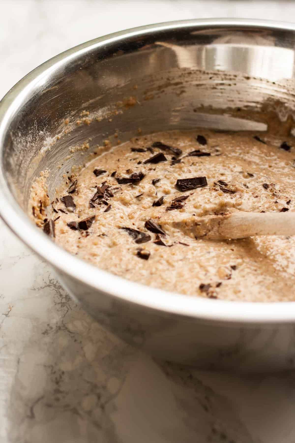 A bowl of muffin batter with a spoon and chocolate chunks inside.