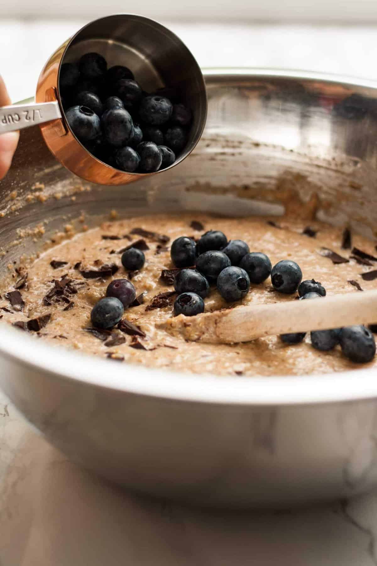 A cup of blueberry being poured into a bowl of muffin batter with a wooden spoon.