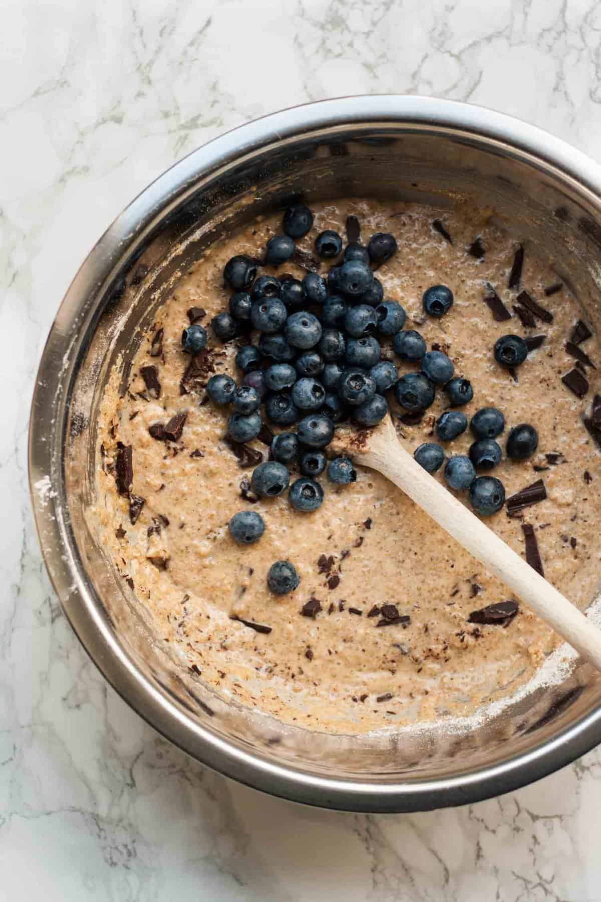 A bowl of muffin batter with blueberries being stirred into it with a wooden spoon.