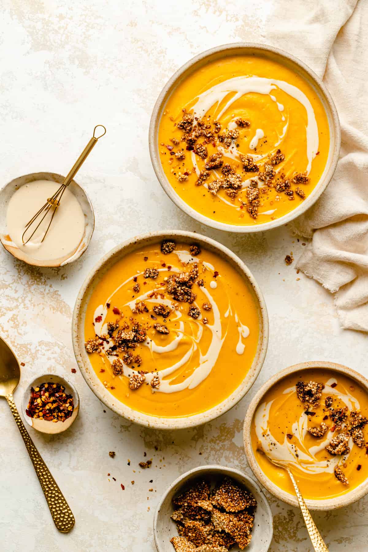 Various bowls of sweet potato soup with gold spoons and side dishes of sesame brittle, chilli flakes and tahini drizzle.