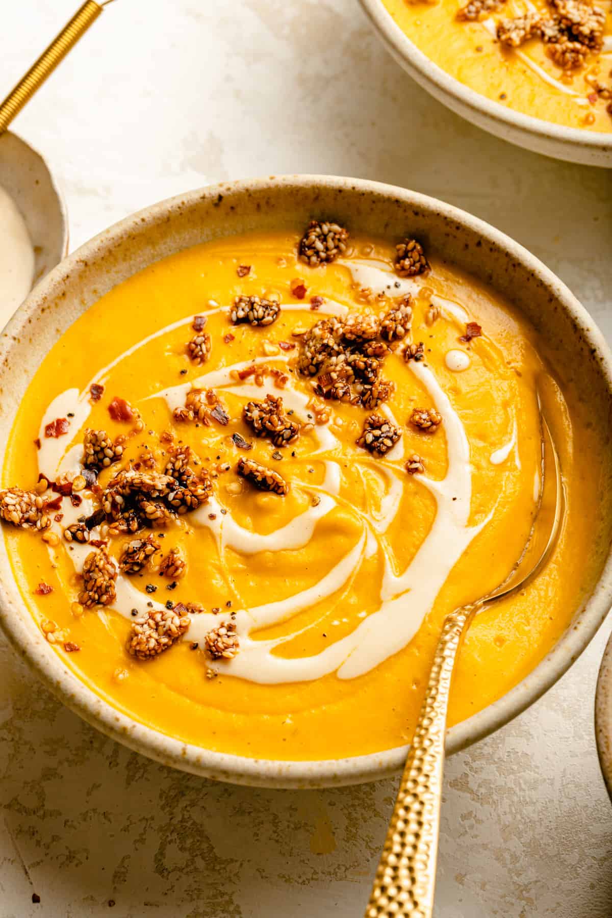 Close up of a bowl of sweet potato and carrot soup with a gold spoon topped with tahini drizzle, sesame brittle and chilli flakes.
