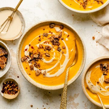 Various bowls of spiced sweet potato and carrot soup with gold spoons and topped with tahini drizzle and sesame brittle.