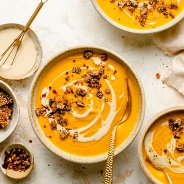 Sweet potato and carrot soup in various bowls with gold spoons, topped with tahini drizzle and sesame brittle.