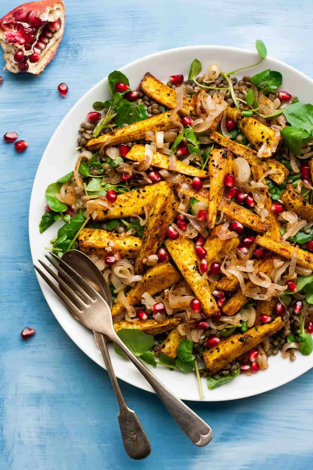 A close up of roasted spiced parsnips on a platter with serving utensils to the side.