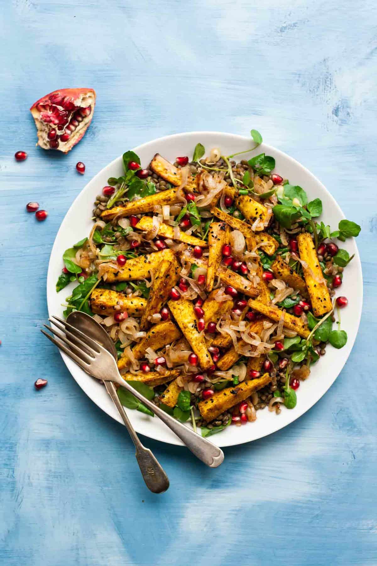 A serving plate with roasted spiced parsnips on top of a lentil salad with utensils.