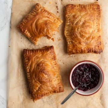 three turkey pastries on parchment with a bowl of cranberry sauce and a bite taken from a pastry.