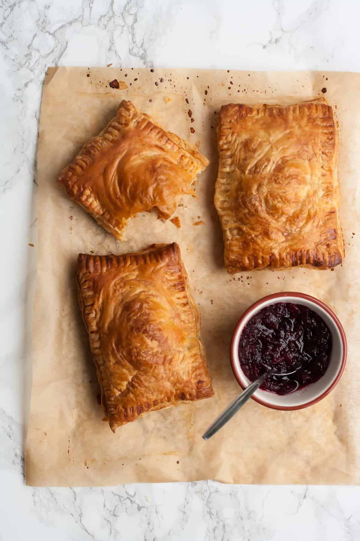 three turkey pastries on parchment with a bowl of cranberry sauce and a bite taken from a pastry.