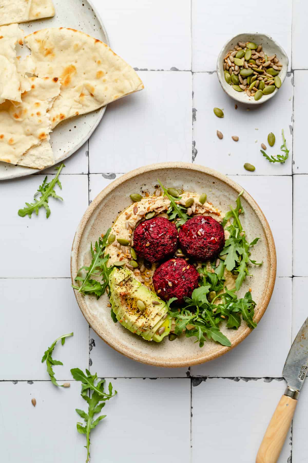 A bowl of beetroot falafel and hummus on a tiled background with a plate of pita on the side.