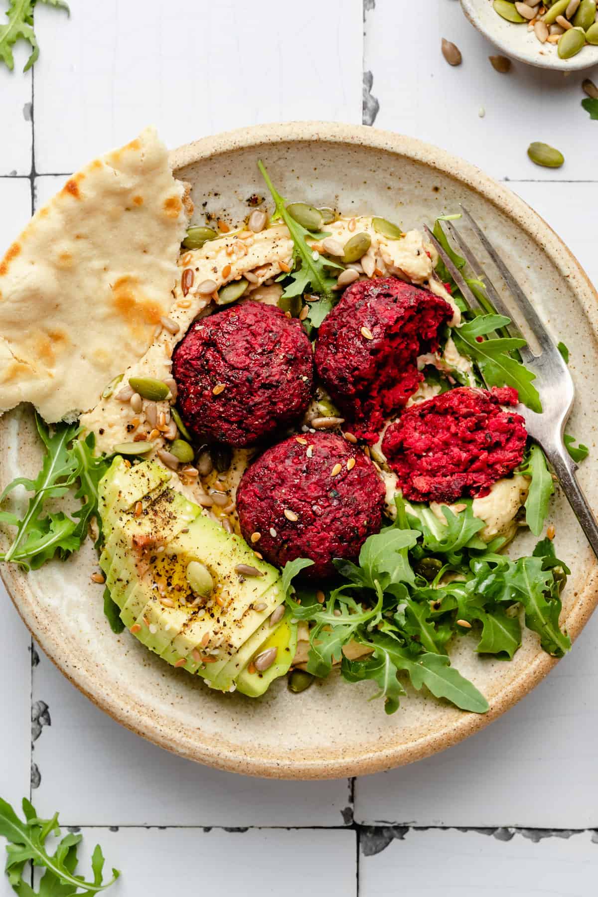 A bowl of beetroot falafels served on top of hummus, with avocado and pita bread, drizzled with oil and seeds.