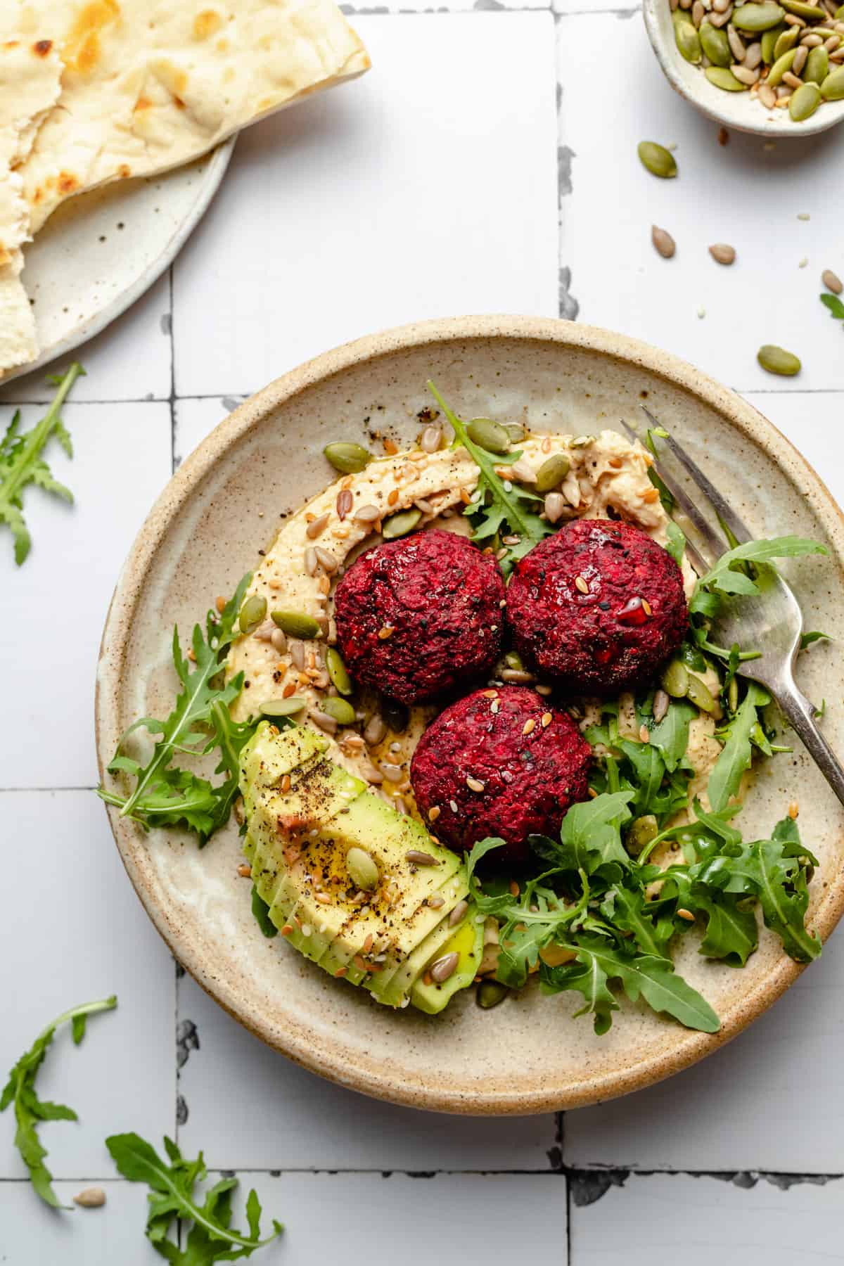 Beetroot falafel served on top of creamy hummus with avocado, seeds, olive oil and arugula, a plate of pita on the side.