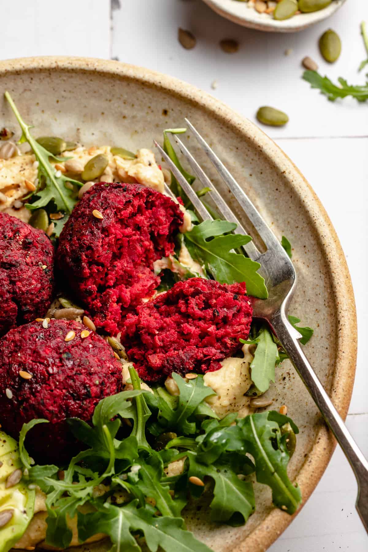 A bowl with broken open beetroot falafel on a bed of hummus with arugula and a fork.