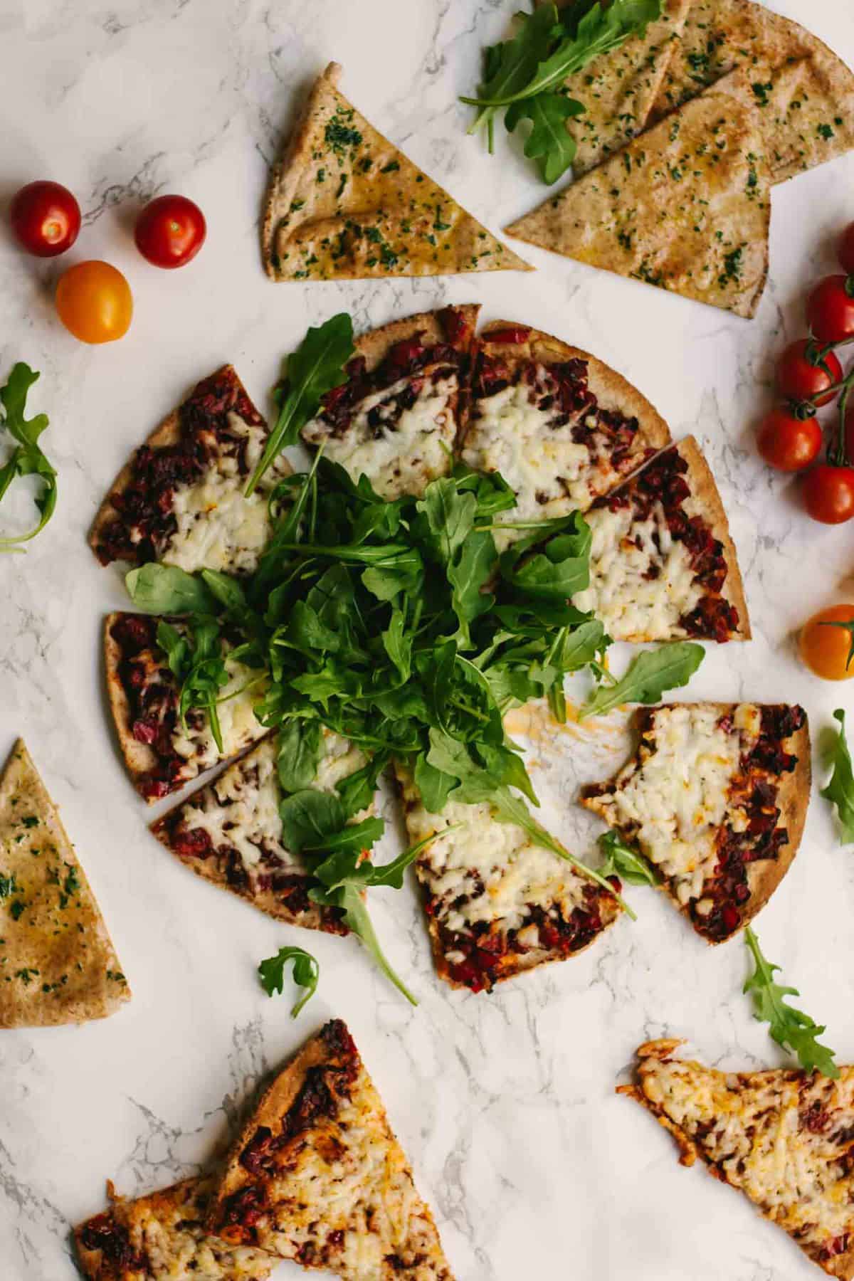 Sliced cheesy flatbread on a marble surface with salad and tomatoes around.