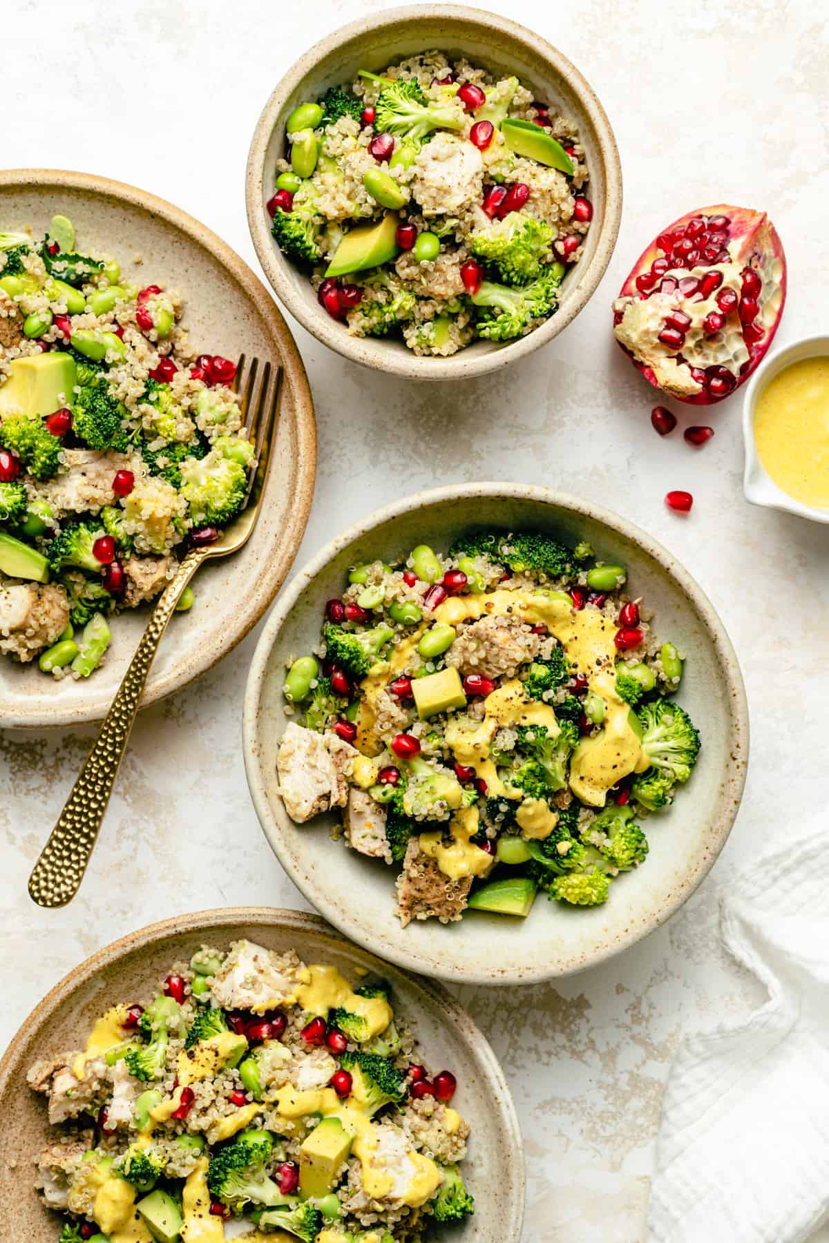 various bowls of chicken quinoa salad with pomegranate arils on top, pomegranate to the side with turmeric dressing in a jug.