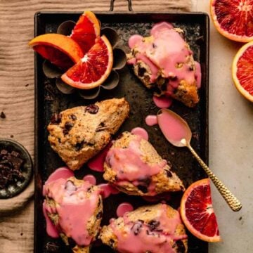 Chocolate chip scones being glazed on a baking sheet with orange slices around with a spoon of glaze.