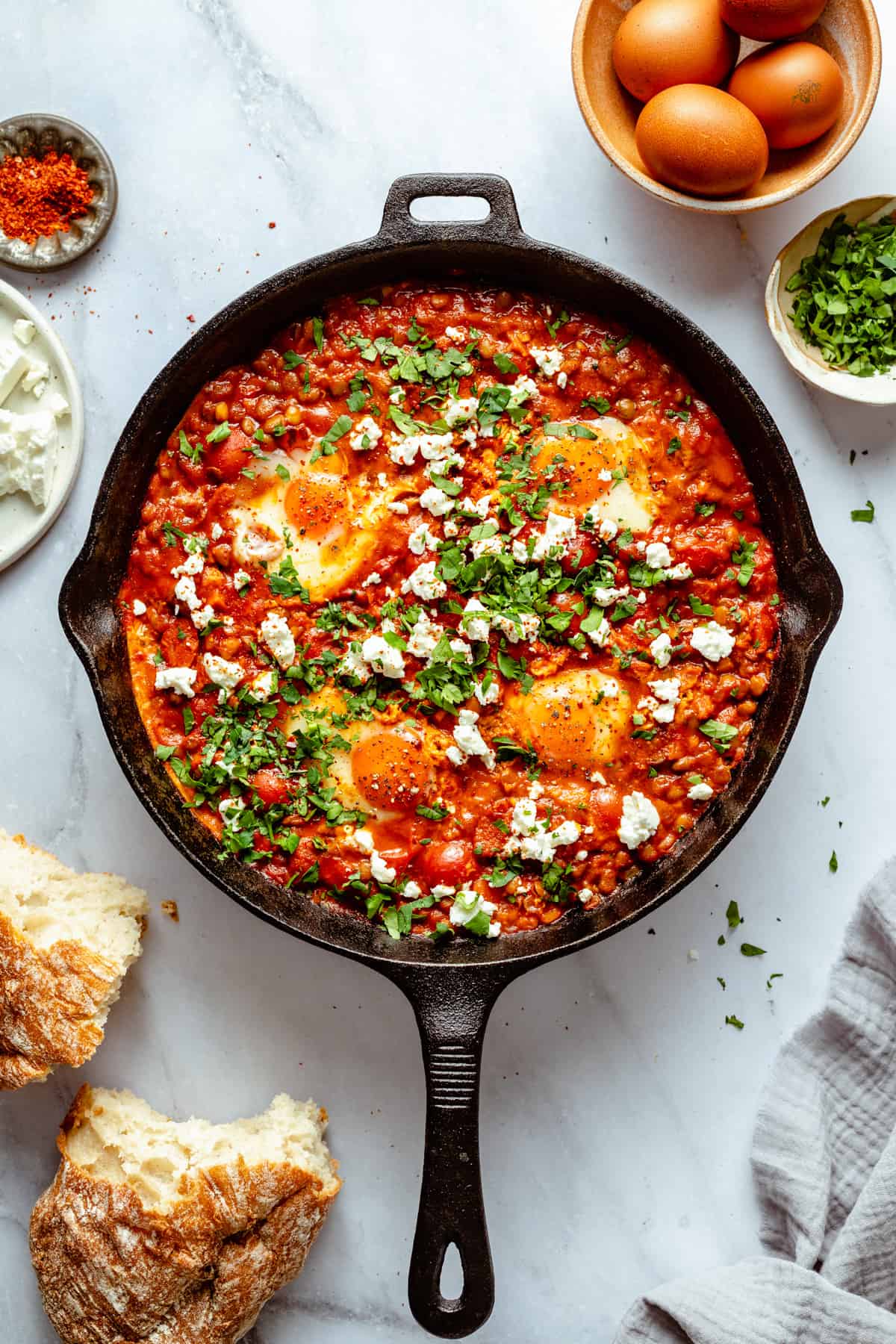 Shakshuka with feta and chorizo in a skillet with bread eggs and herbs around.