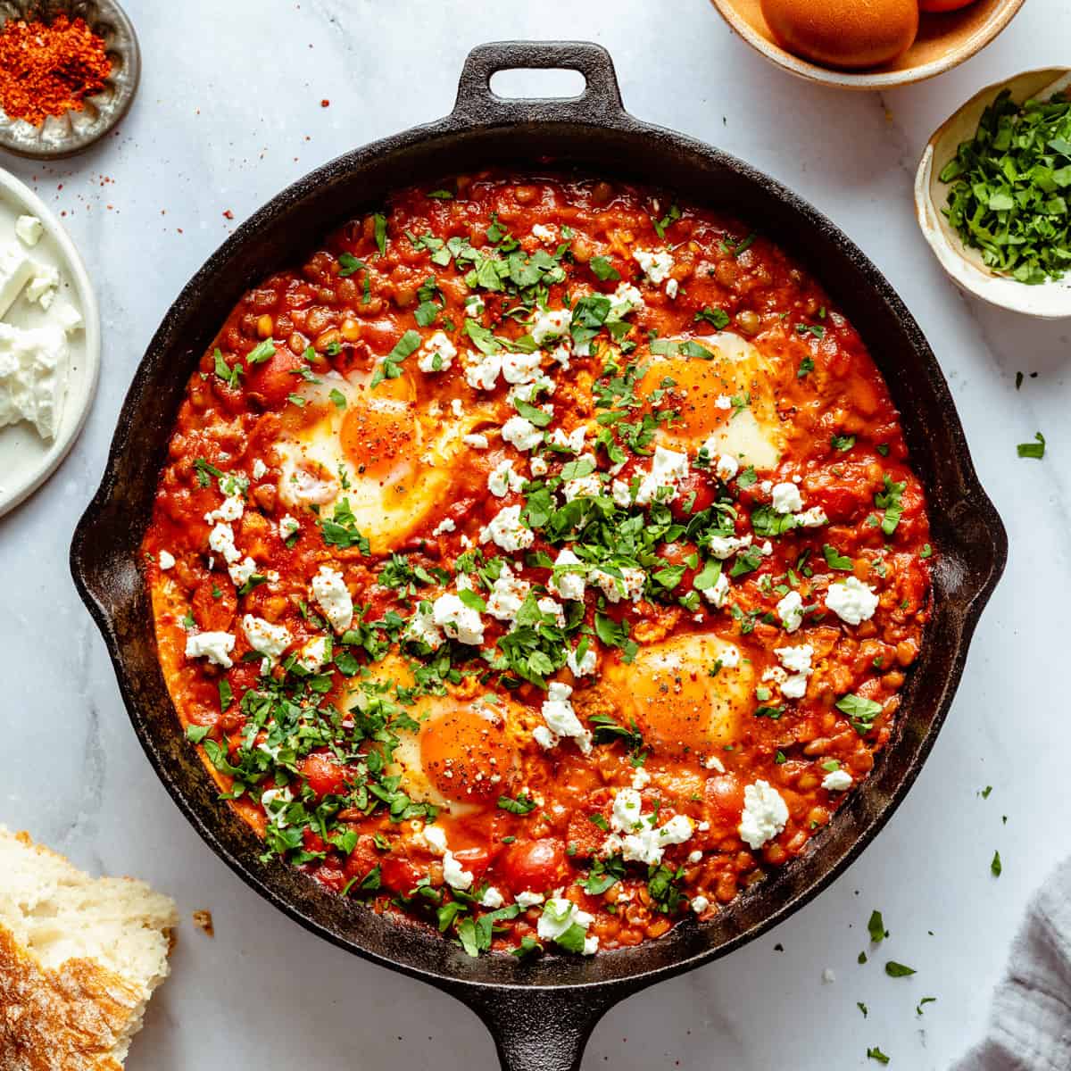 Shakshuka with feta and chorizo in a skillet with herbs, eggs and bread around it. 